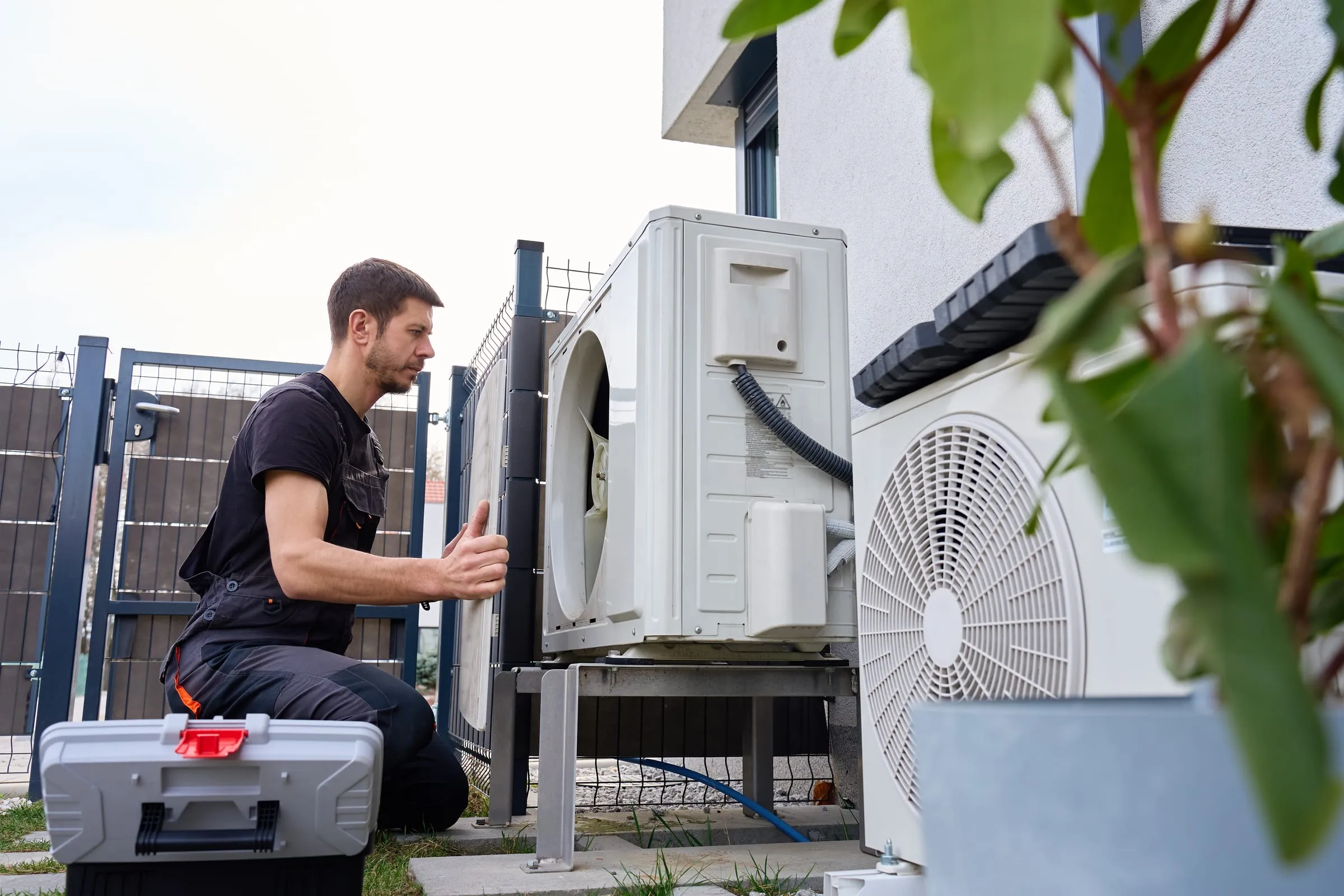 Technicien intervenant sur l’unité extérieure d’une pompe à chaleur, caisse à outils au sol.